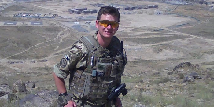 Man in military jacket and sunglasses standing in front of a mountainous desert landscape