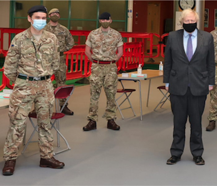 Military and government personnel in PPE and face masks standing near red barriers at an official engagement